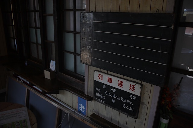 折居駅の駅舎内に残る、木製の古い黒板と「列車遅延」の案内板。黒板の左端には「西浜」「折居」「三隅」といった駅名が手書きされており、時代を感じさせるノスタルジックな風景。