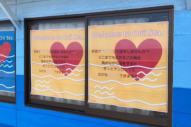 折居駅の青い駅舎の窓に貼られた、大きな赤いハートが描かれたポスター。「折居でおりいった話をしませんか?」という駅名にかけたメッセージが書かれている。