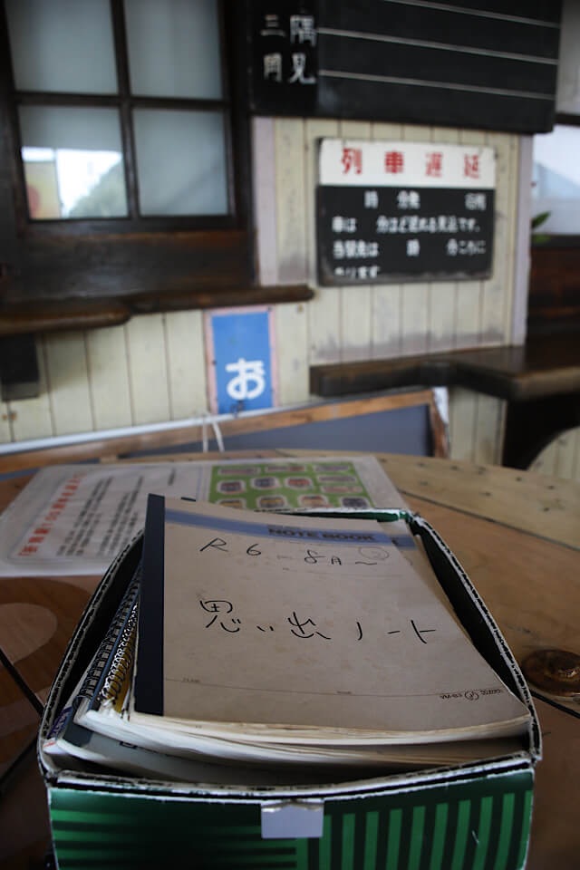 折居駅の待合室に置かれた、旅人のための「思い出ノート」。緑色の箱の中にノートが重なっており、表紙には「R6 8月〜」「思い出ノート」と手書きされている。背景には駅舎のレトロな内装がぼけて映り込んでいる。
