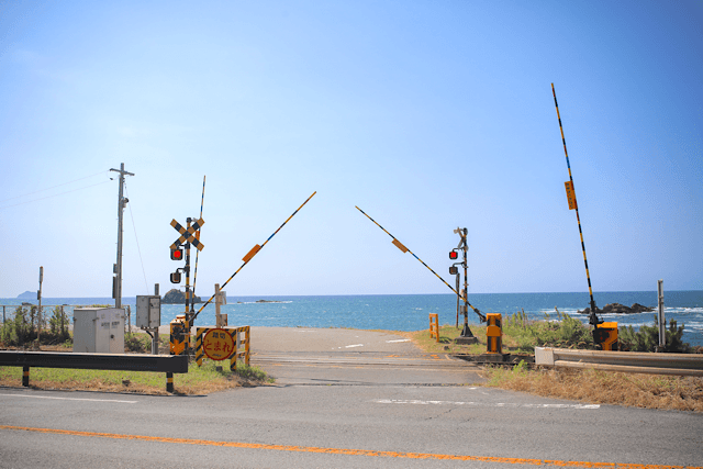 折居駅近くの「山陰街道踏切」。上がりかけた遮断機の向こう側に、水平線まで一面に広がる青い日本海と快晴の空が見える。