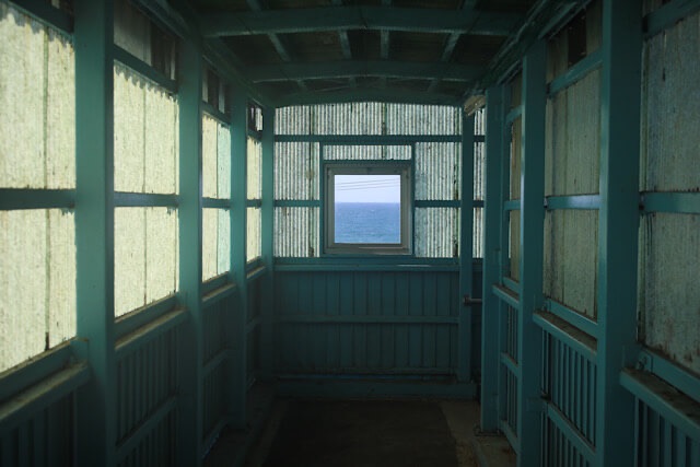 折居駅の、時の流れを感じさせるレトロな跨線橋内。古びた窓枠が中心にあり、その向こう側に青空と深い青色の日本海を完璧に切り取った風景が見える。