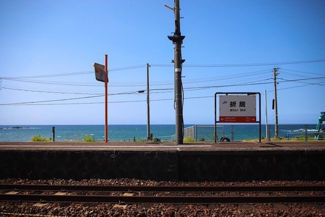 島根県にあるJR山陰本線・折居駅のホーム。中央に立つ「折居駅」の駅名標の向こう側に、水平線が広がり、真っ青な日本海と快晴の空が広がっている風景。