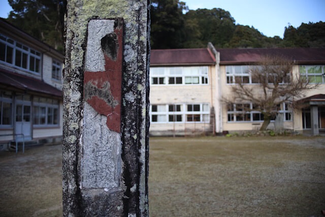 手前に大きく映し出された、文字が剥げかかった「九鬼小学校」の古い門柱。背景には、ぼんやりと静かに佇む木造校舎と校庭が写っている。