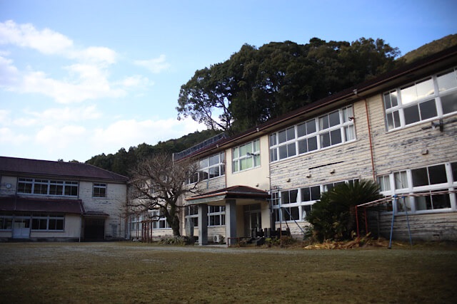 青空と緑の山を背景に、静かに佇む旧九鬼小学校の木造校舎。誰もいない広い校庭と、歴史を感じさせる重厚な建築美。