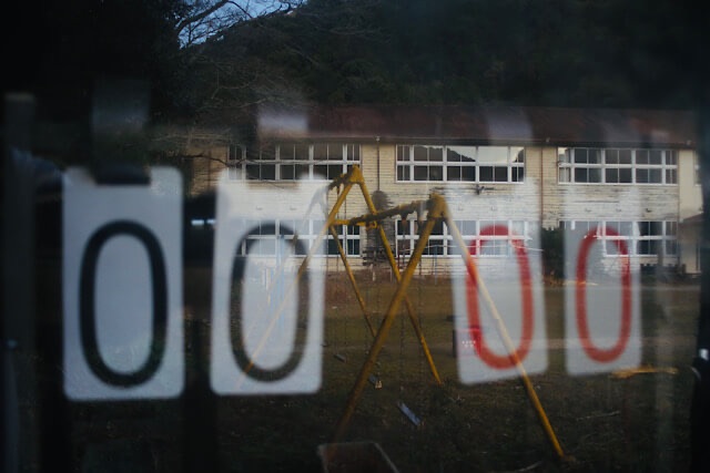倉庫のガラスに映る、旧九鬼小学校の校庭と木造校舎。倉庫の中には古いスコアボードが残されており、数字「0 0 0 0」が表示されている。奥には草に埋もれかけた錆びたブランコが立っている。