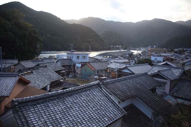 高台（旧九鬼小学校付近）から見下ろす九鬼町の全景。手前に密集して並ぶグレーの瓦屋根の家々、その奥に穏やかな湾、そして背後を囲む深い山々が写っている。