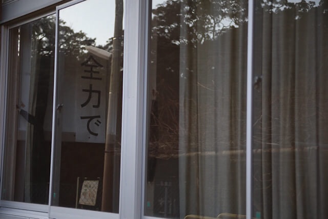 窓ガラス越しに見える、「全力で。」と縦書きで力強く書かれた校内掲示。ガラスには外の木々が重なるように反射しており、閉ざされた教室の静寂を感じさせる。