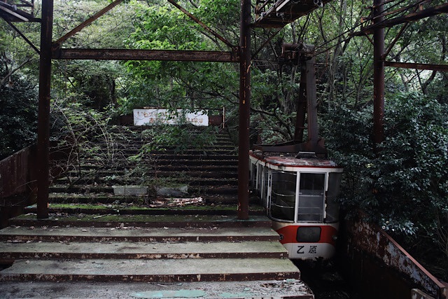 志高ロープウェイ立石山駅のホーム跡。左側の苔むした階段の先に「いらっしゃいませ ありがとうございます」の看板が見え、その右側で「乙原」と書かれた赤白のゴンドラが地面へと沈み込むように静止している。全体を錆びた鉄骨と深い緑が覆っている。