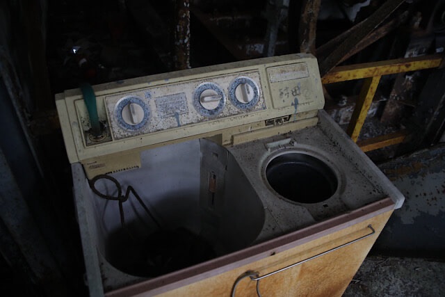 暗い廃墟の機械室の床に、古びてプラスチックが黄ばんだ二槽式洗濯機が置かれている。錆びたダイヤルや金属パネル、洗濯槽と脱水槽が見える。背景には錆びた鉄製の機械がぼんやりと写っている。