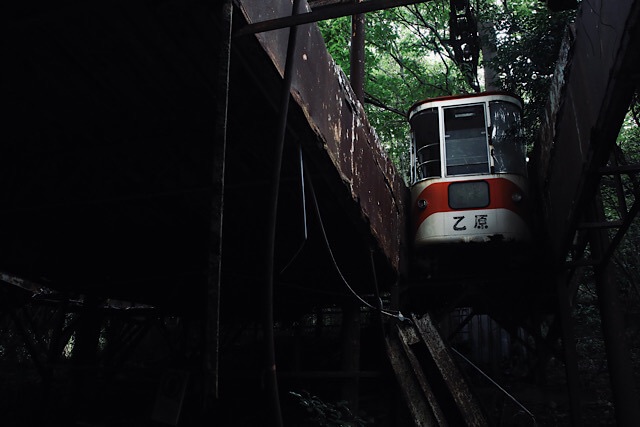 暗い森の廃墟（立石山駅舎）の中に、錆びついた鉄骨構造物と、そこに残された赤と白の志高ロープウェイのゴンドラ。ゴンドラの車体には「乙原」と書かれている。