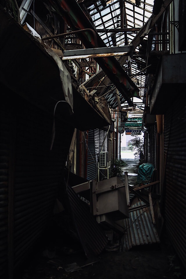 崩壊が進む下渕マーケットのアーケード内部。錆びたシャッターが続く暗い通路に、破損した屋根から光が差し込み、崩落した瓦礫（トタンや構造物の残骸）が散乱して通路を塞いでいる。瓦礫の先には明るい出口と「喫茶 クワヤマ」の看板が見える。