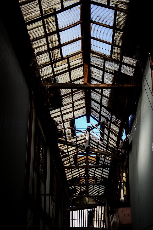 廃商店街「下渕マーケット」のアーケード内部で、崩壊した天井屋根を見上げた写真。波板の屋根が大きく破損し、露出した古い木造の梁の隙間から、青空がはっきりと見えている。左右の暗い壁面と、奥に見える明るい出口が対比されている。