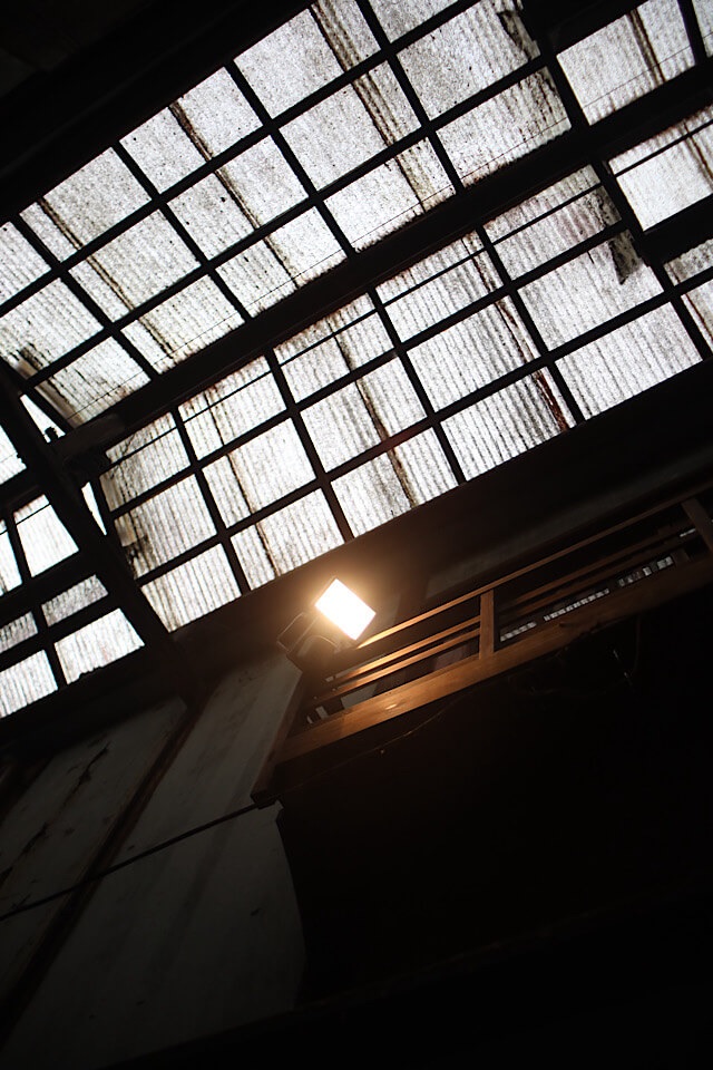 崩落が進む下渕マーケットの天井付近、格子状の波板屋根を背景に点灯している人感センサーライト。暗い建物内部でライトだけが明るく光り、周囲の劣化した柱や屋根の質感を浮かび上がらせている。