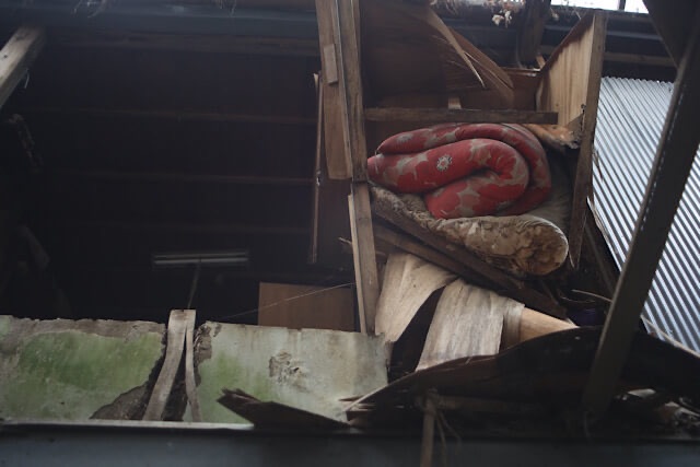 崩壊が進む下渕マーケットの廃墟で、朽ちかけた木造構造物の間に赤い地に白い模様の布団（毛布）が挟まっている。周囲の壁は緑色に風化し、劣化が進んでいる。