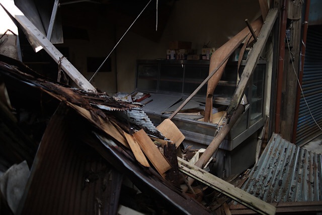 崩壊した下渕マーケットの店舗内部。激しく歪んだトタンや木材の瓦礫が山積みになり、通路を完全に塞いでいる。奥には、かつて商品を陳列していたと思われるガラスのショーケースが無残に残されている。