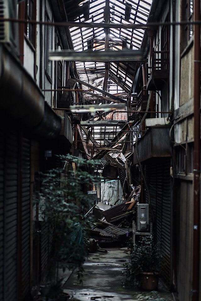 下渕マーケットのアーケード内部を入り口から奥へと見通した写真。暗い通路の先は、崩落した天井や建物の残骸が山積みになって完全に塞がれている。格子状に残った屋根の骨組みから冷ややかな光が差し込み、手前には放置された植木が自生している。