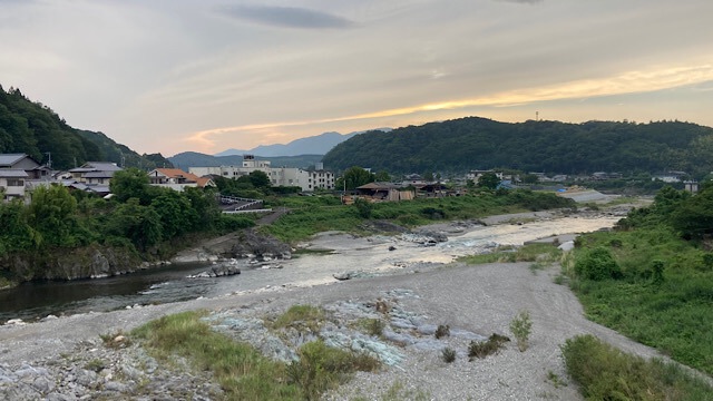 下渕マーケットのすぐ裏手を流れる吉野川の広大な風景。夕暮れ時の淡い光の下、穏やかに流れる川と広い河原、背景には緑豊かな山々と静かな町並みが広がっている。