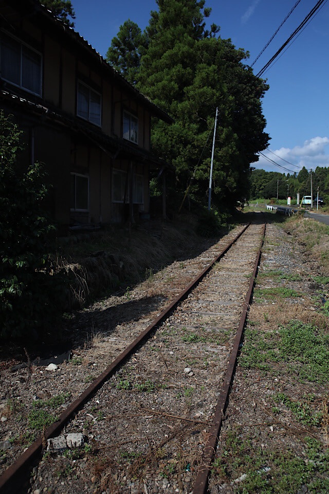 住宅のすぐ脇をまっすぐに伸びる旧国鉄倉吉線の廃線跡。錆びた2本のレールが露出し、そのすぐ左手には生活を感じさせる古い木造民家が建っている。空は青く、レールの周りのバラストも比較的きれいに残っている様子。