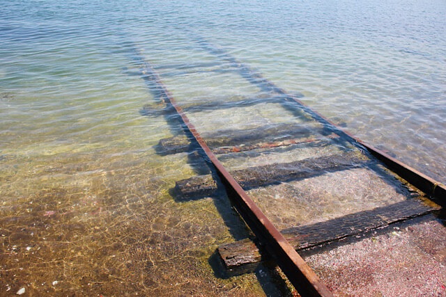 房前漁港の海へと続く線路を斜めのアングルから捉えた写真。透明な瀬戸内海の水面下に沈んでいく錆びたレールと木製の枕木が、陽光に照らされて美しく輝いている。静かでノスタルジックな春の風景。