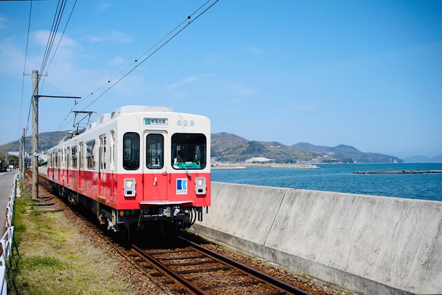 瀬戸内海のすぐ脇を走る琴電志度線の赤と白のレトロな車両。青空と穏やかな海、遠くの島々を背景に、単線の線路をゆっくりと走行する様子。