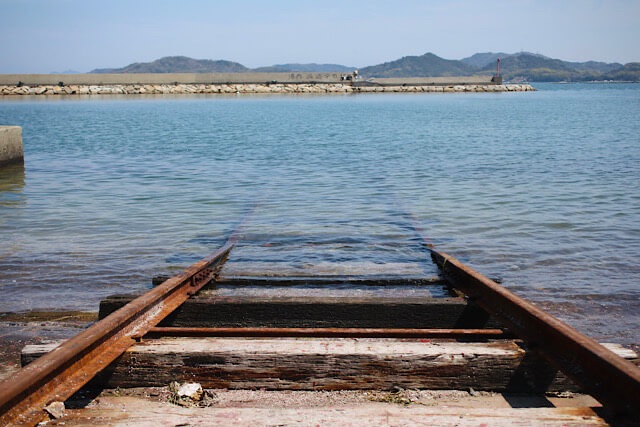 香川・房前漁港の海中線路をローアングルから捉えた写真。錆びたレールと使い込まれた枕木が透明度の高い海へと吸い込まれていく様子が、奥行きを持って写っている。遠景には穏やかな海と防波堤、山々が広がる。