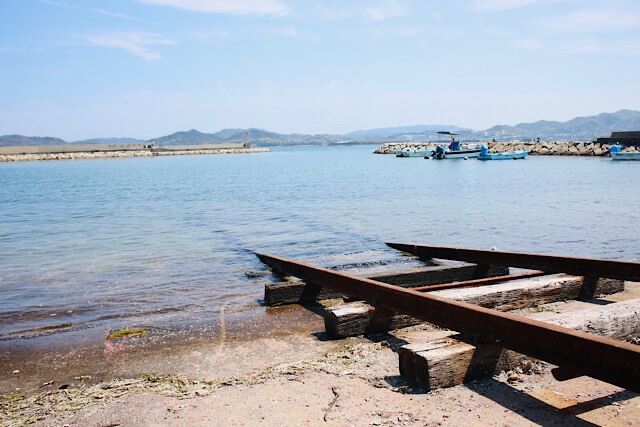 香川県房前漁港の海へと続く線路（インクライン）の横構図写真。陸上の錆びたレールと枕木が、透明度の高い穏やかな瀬戸内海へと伸び、そのまま水中に沈んでいる。水中の線路もはっきりと見え、遠景には港と山々が広がる。