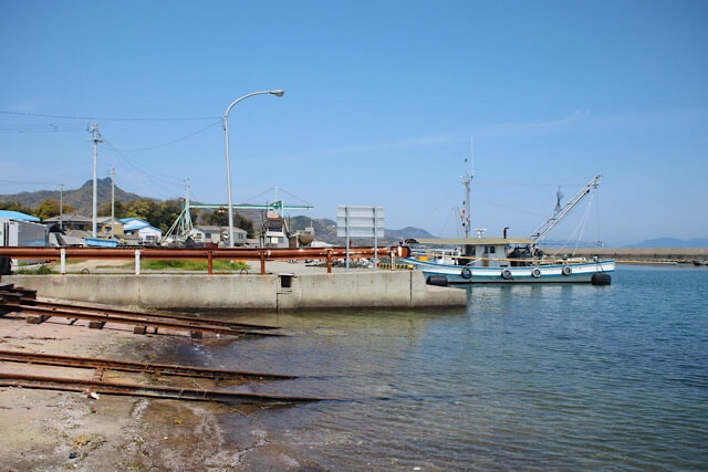 房前漁港の「海へと続く線路」を少し引きの構図で捉えた写真。錆びたレールが波打ち際から海中へと伸び、右側には白い漁船が停泊している。晴天の下、穏やかな瀬戸内海と遠くの山々が広がる静かな風景。