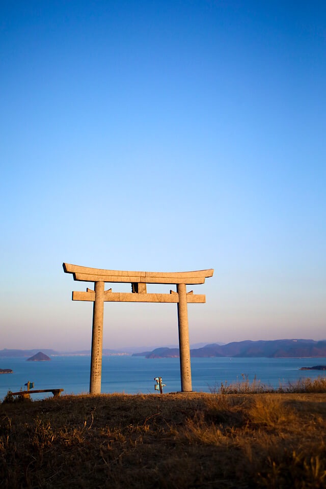 龍王山頂上付近の天空の鳥居。夕陽の光を浴びてオレンジ色に輝く石造りの鳥居と、その向こうに広がる瀬戸内海の多島美、澄んだ青空。