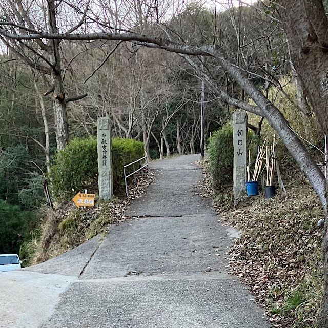 龍王山ハイキングのスタート地点。「朝日山聖観世音菩薩」の文字が刻まれた石柱と、木々に囲まれた舗装された上り坂の入り口。