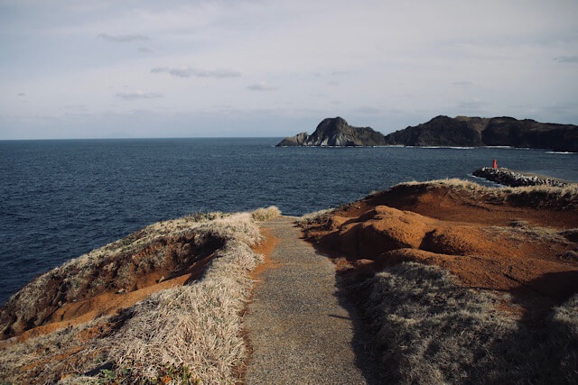 見島の赤い岩肌が広がる断崖から、深い青色の海へと続く砂利道。遠くには島影が浮かび、右側の突堤には小さな赤い灯台が見えるダイナミックな風景。