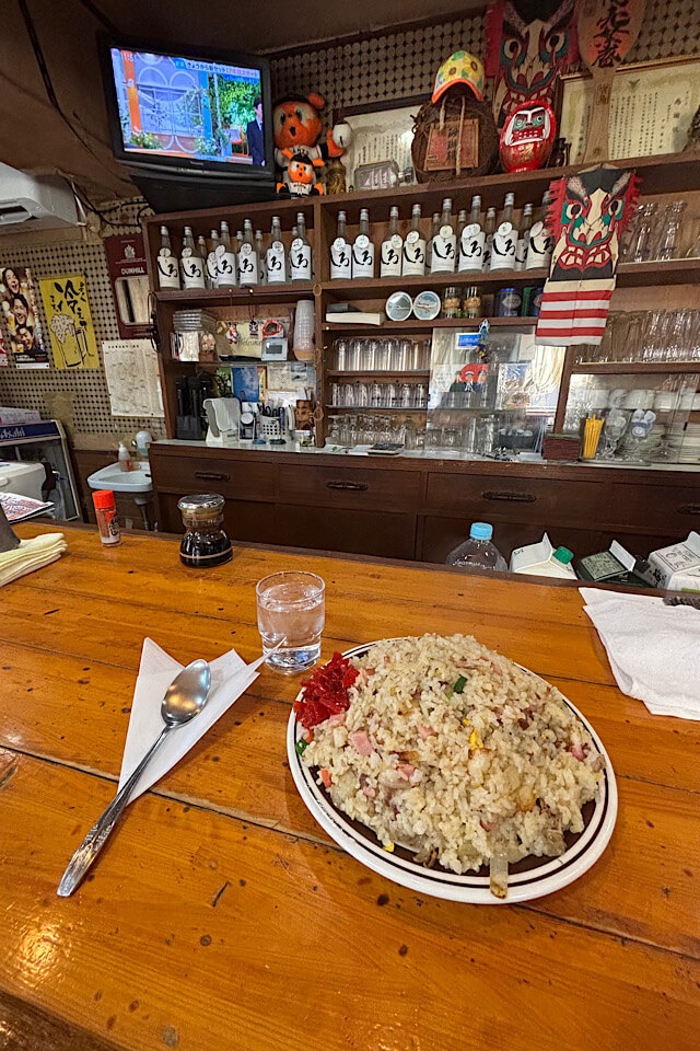 木製のカウンターに置かれた、山盛りのチャーハンの皿。奥には、焼酎の瓶、テレビ、様々なお守りや装飾品が並ぶ、懐かしい雰囲気の店内が見える。