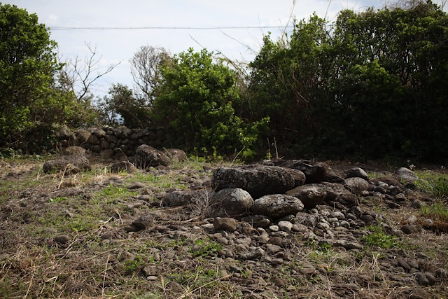 ゴロゴロとした石を積み上げた古墳が、荒れた草地、砂利、点在する岩の中に立っている。背景には、曇り空の下、青々とした木々、石垣、裸の枝が見える。