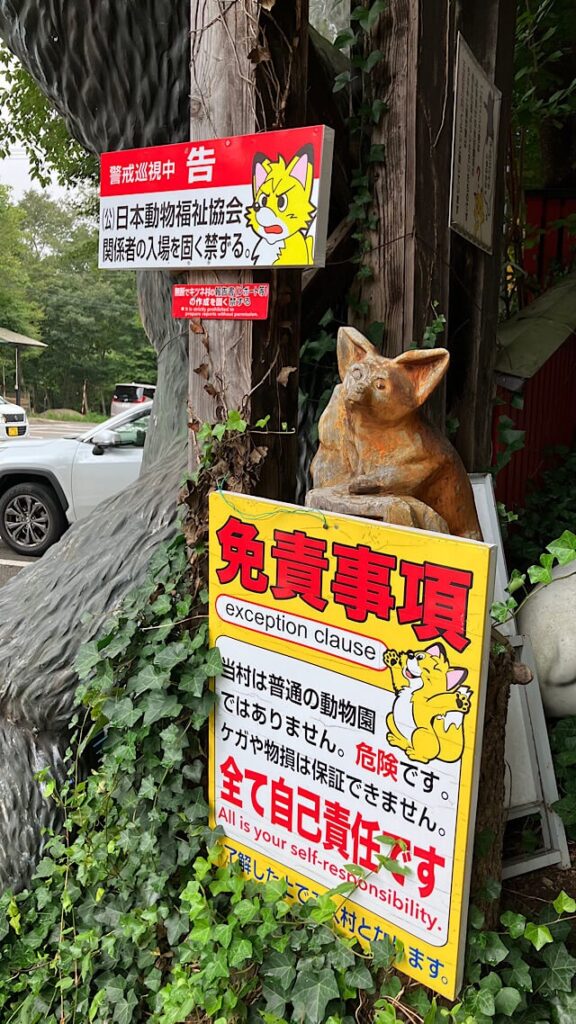蔵王キツネ村の入り口に設置された複数の警告看板。上部の赤と白の看板には「(公)日本動物福祉協会関係者の入場を固く禁ずる」とあり、下部の黄色い看板は「免責事項」で、「当村は普通の動物園ではありません。危険です。ケガや物損は保証できません。全て自己責任です」との警告文と、その下に「All is your self-responsibility」の英語表記。看板の後ろには木製のキツネ像があり、背景に木々や白い車が見える。
