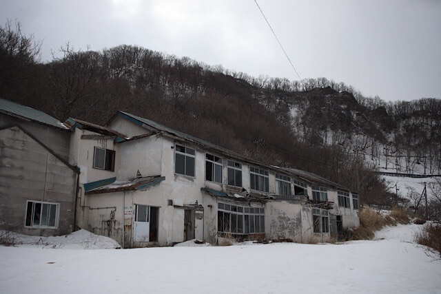 雪が積もった冬の岩山を背景に、雪原の中にポツンと現存する雷電温泉「ホテル観光かとう」の白い建物全景。窓ガラスが一部割れ、老朽化が進んでいる廃墟の外観。