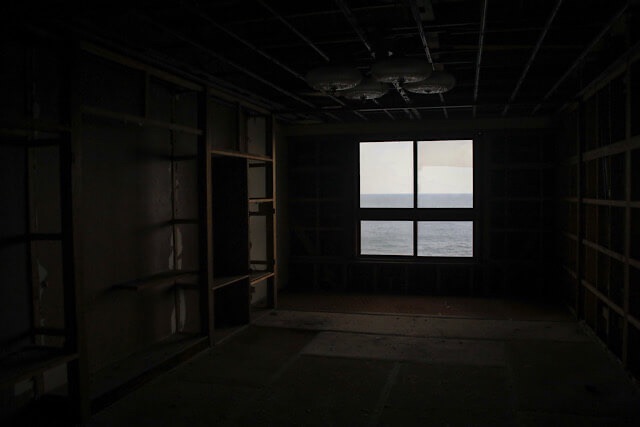 雷電温泉「ホテル観光かとう」の廃墟となった客室内部。壁や天井の仕上げが剥がれ落ちて格子状の骨組みが剥き出しになった薄暗い部屋の奥に、日本海が見える四角い窓がある光景。