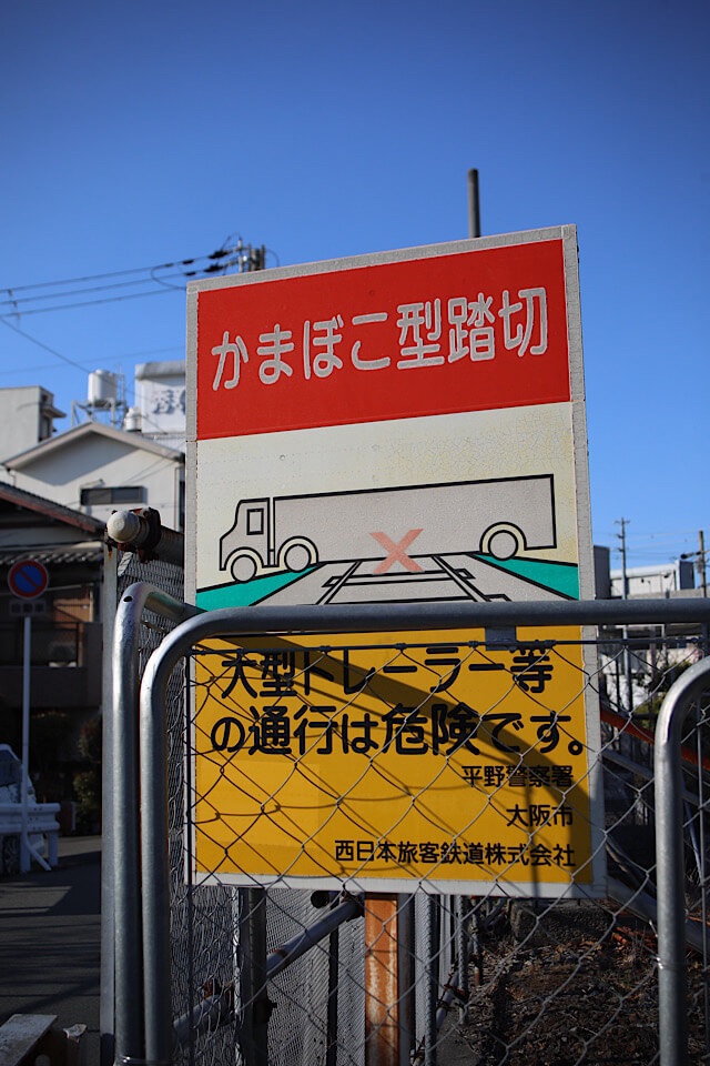 フェンス越しに見える、平野警察署・大阪市・西日本旅客鉄道株式会社が設置した「かまぼこ型踏切 大型トレーラー等の通行は危険です。」と書かれた警告看板