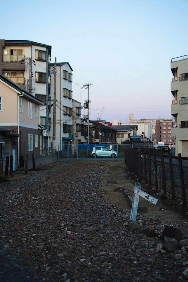 立ち並ぶマンションや住宅の合間に広がる砂利の空き地と、手前に残された「4.0」と書かれたコンクリート製の鉄道用距離標