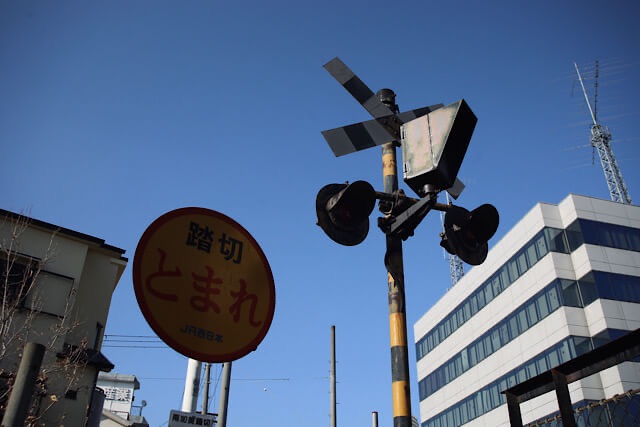 青空を背景に、警報灯やバツ印の検知器、JR西日本の「踏切とまれ」の丸型標識が残されている阪和貨物線の踏切遺構