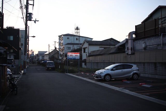 夕暮れの住宅街に馴染むように配置されたコインパーキングと、その奥へと細長く一列に並ぶ建物に挟まれた阪和貨物線の跡地