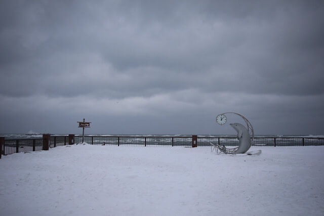 一面の雪で覆われたノシャップ岬の広場。どんよりとした曇り空と寒々しい冬の海を背景に、イルカの時計モニュメントとノシャップ岬の標識が静かに佇む風景。