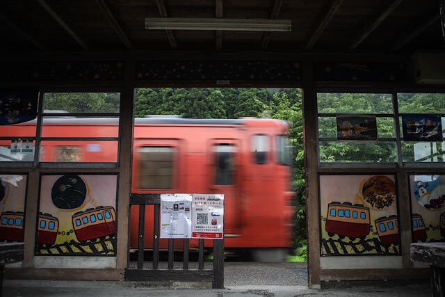飯給駅の木造待合所の中から見る、ホームを勢いよく通過していく赤い気動車の流し撮り風景。