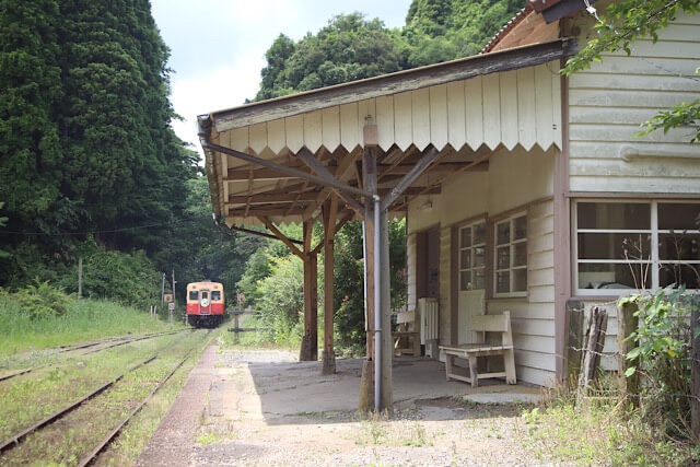 初夏の緑に囲まれた小湊鐵道・月崎駅の木造駅舎とホーム、線路の向こうから入線する赤い気動車。