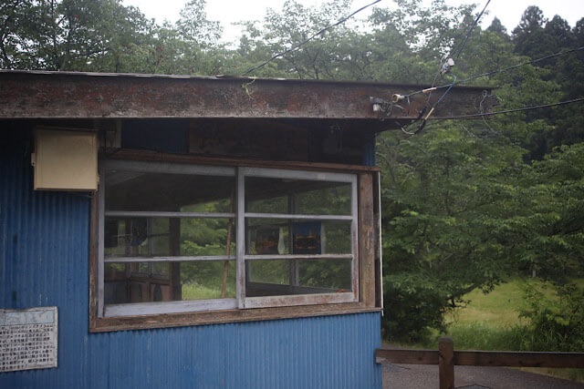飯給駅の青い波トタンと木枠の素朴な待合所と、周囲に広がる里山の緑。